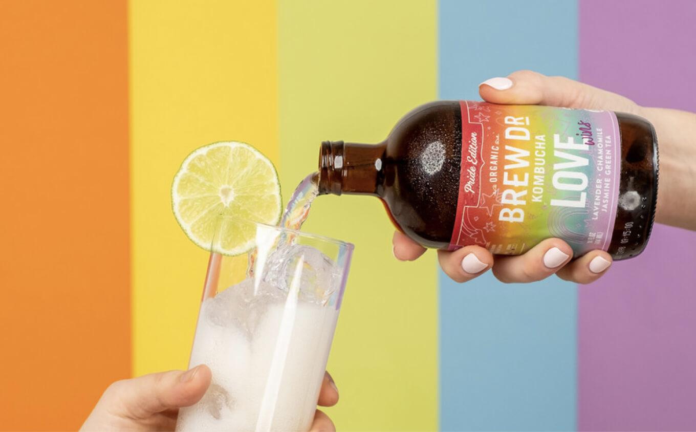 Glass bottle of Brew Dr Kombucha's Pride edition being poured into a glass against a colorful background